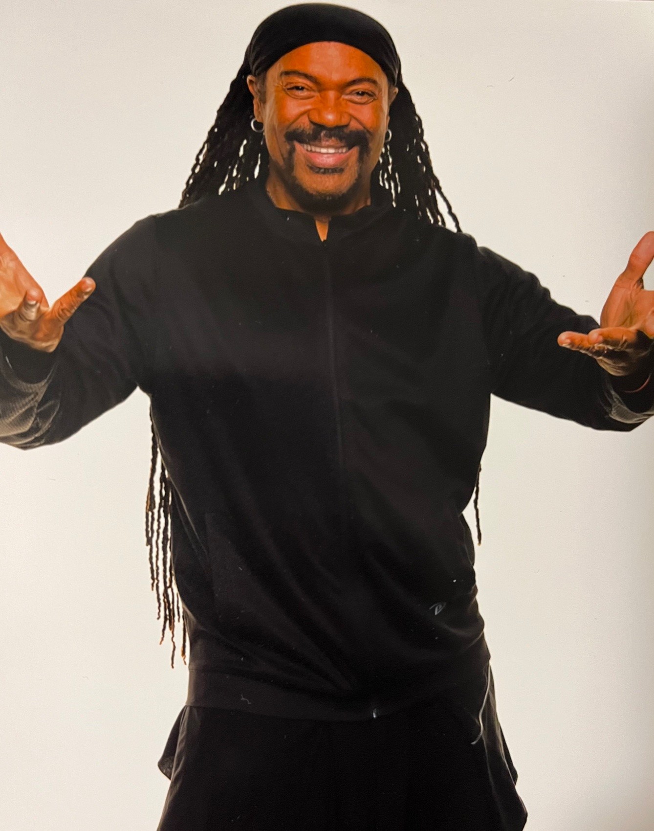 Photo of smiling brown skinned man with waist length locs standing in front of a cream colored background with arms outstretched