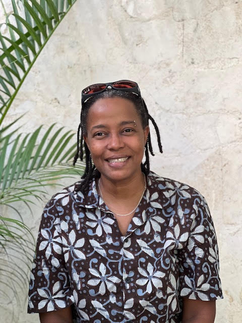 woman with cornrows smiling  in front of a taupe wall wearing a shirt with white flowers
